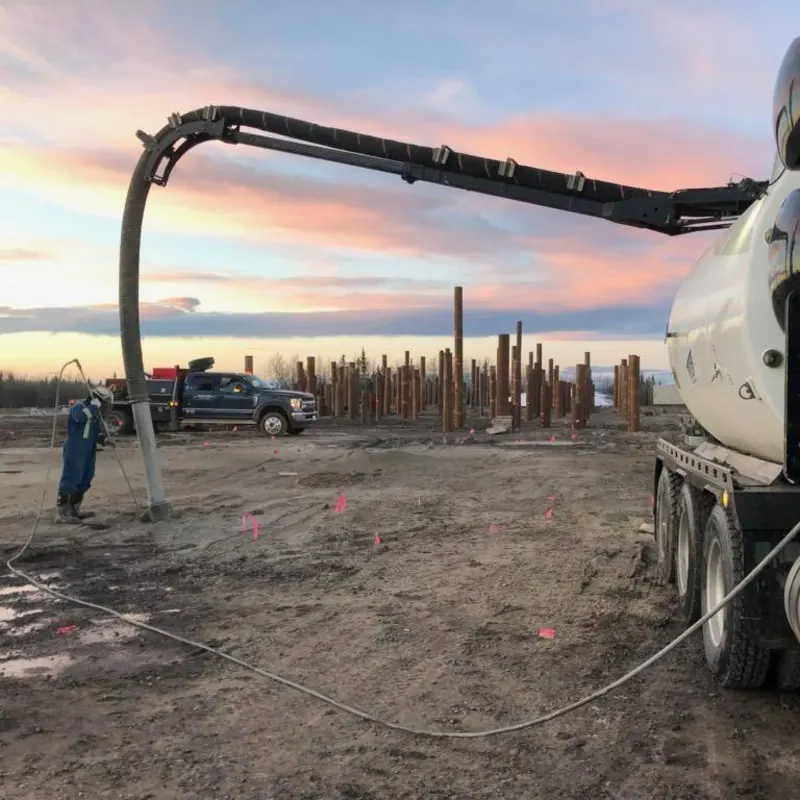 A vac truck boom reaching down to a worker at a site with rows of steel piles at dawn