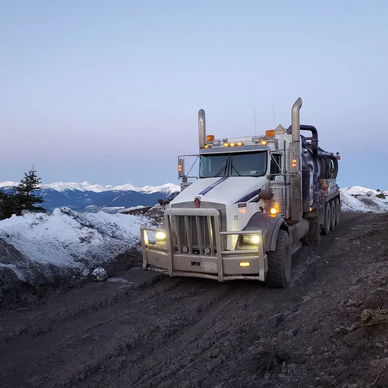 A Troyer vac truck on a snowy mountain road at dusk with its headlights on