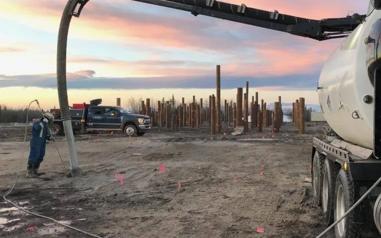 A Troyer vac truck boom reaching down to a worker at a site with rows of steel piles at dawn
